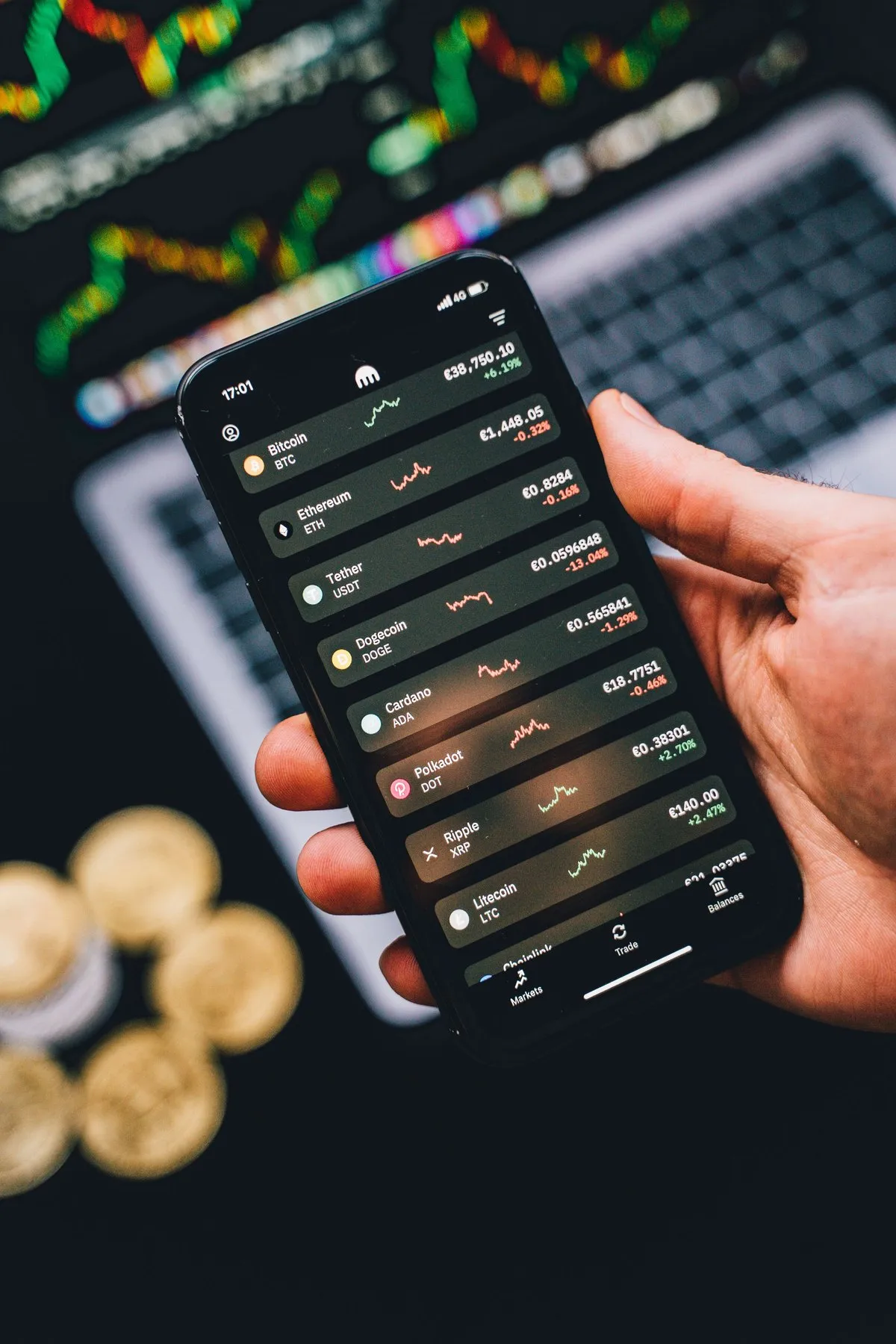Hand holding a smartphone with a crypto trading app above gold coins and a chart screen