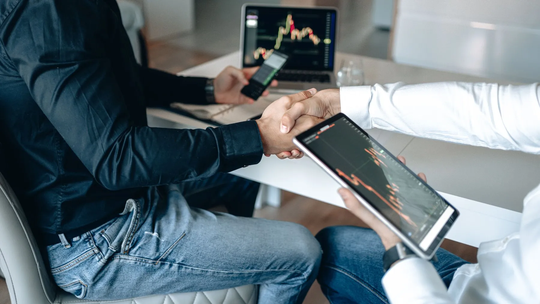 Close-up of a handshake above a tablet showing a crypto price chart