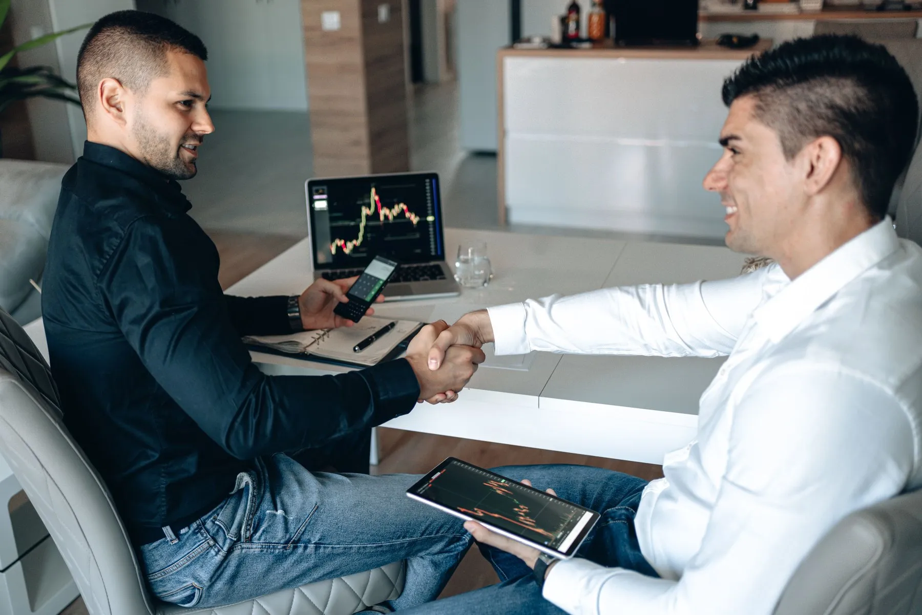 Two business partners shaking hands over a desk with trading charts on a laptop and tablet